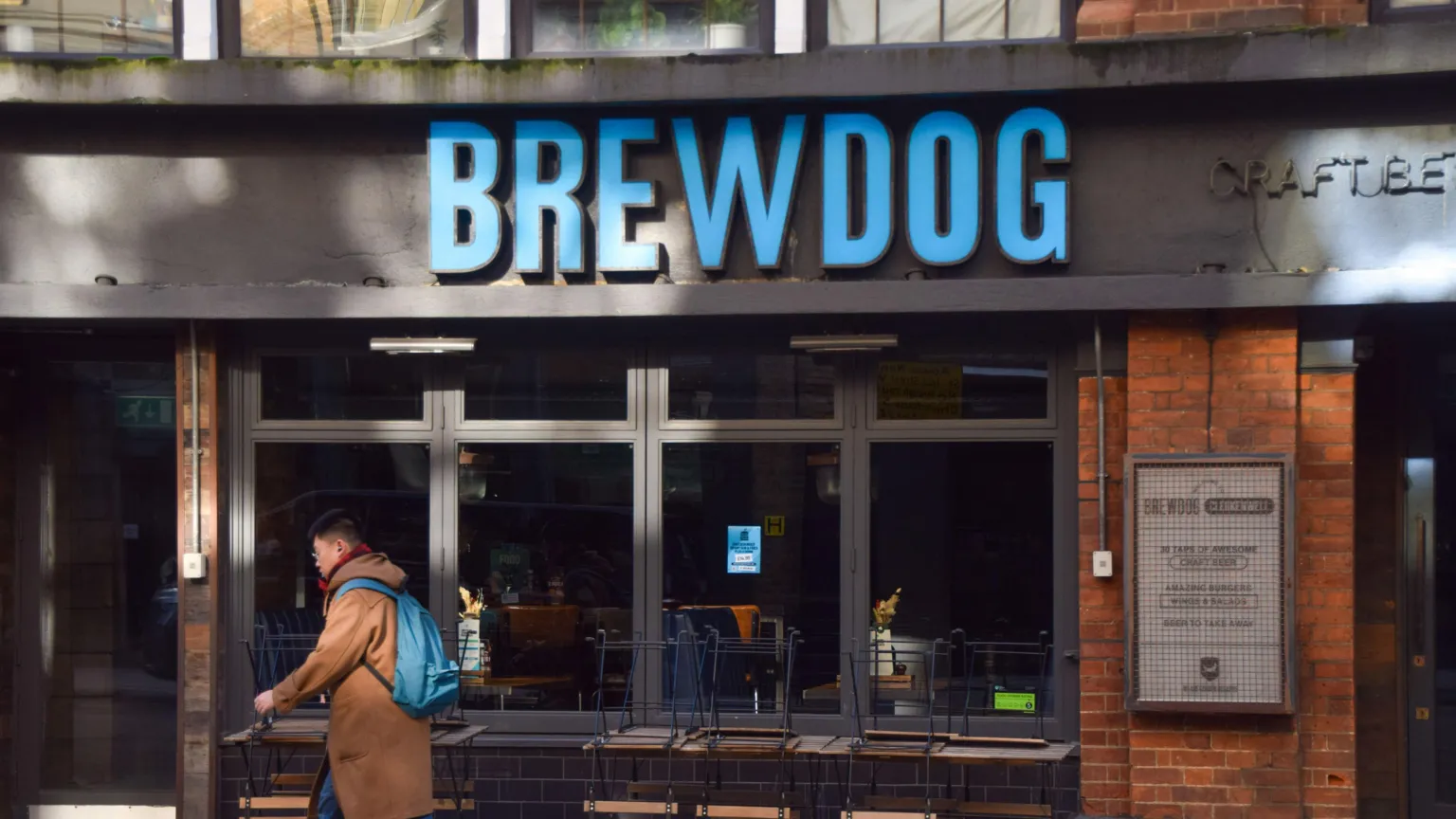 A person walks past a Brewdog bar. The sign on the wall has the name of the company in large blue letters.