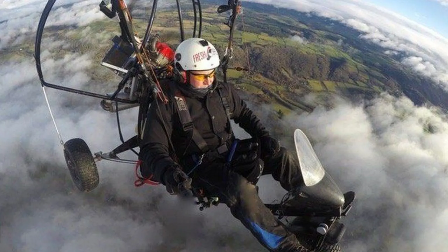 Conservation Without Borders Dan Burton is in the sky in a light aircraft called a paramotor. There is no cockpit and Mr Burton, who is wearing a white helmet and black flying suit, is exposed to the elements. The aircraft has a large motor behind the pilot. Mr Burton is flying above clouds and green fields.