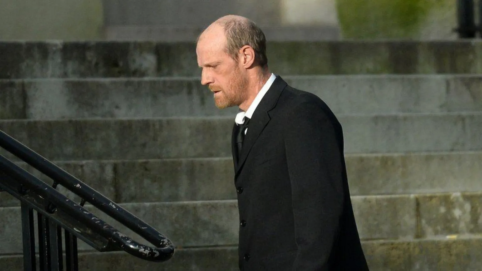 Wales News Service A man is walking to some steps, wearing a black wool coat, a white shirt and a black tie. Behind him are the dirty stone walls of the court building.