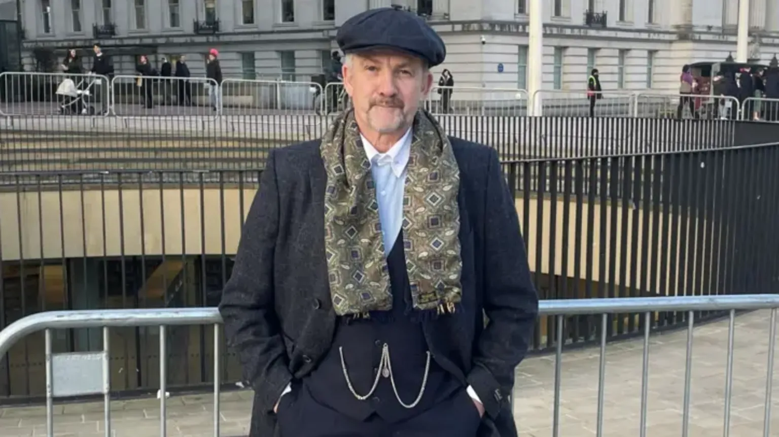 Arthur Cocker, a man dressed in a dark-coloured waistcoast and jacket with a white shirt and a dark-coloured flat cap with a patterned scarf. He is standing by railings in a city centre.