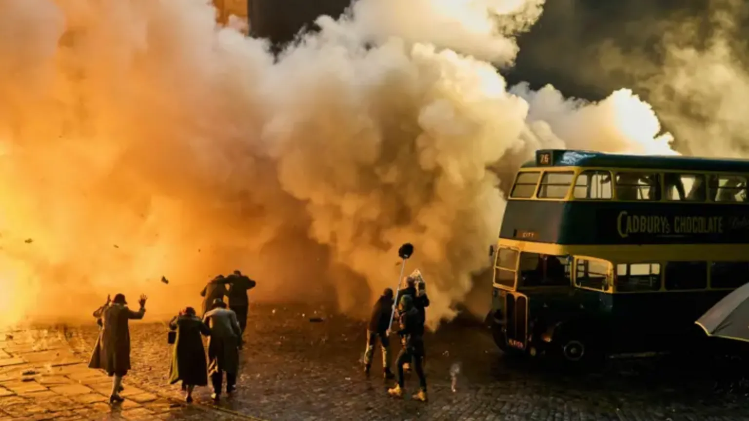 Netflix A film set featuring a group of actors and film crew standing in front of a double decker bus, as an explosion engulfs much of the image. Many of the actors appear to be holding their hands up or shielding themselves from the blast.