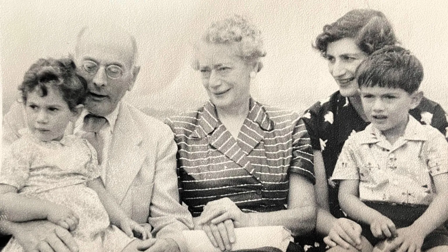 Moritz Family Photo A black and white image of the Moritz family. Sir Michael is a child in this image and is pictured on the far right sitting on his mothers knee, he has dark hair and a white patterned shirt on, as well as dark shorts. His mother looks to the left of the image and is wearing a black and white patterned dress. She too has dark hair. In the middle is Sir Michael's grandmother, who has a black and white striped dress, her hands are atop a white purse and she has short, white, curly hair. Next to her is a man, Sir Michael's grandfather, who is bald and has round-framed glasses on. He has a white suit jacket, over a white shirt and a lightly coloured tie. He has a baby girl on his lap who has a light-coloured dress on and dark curly hair.