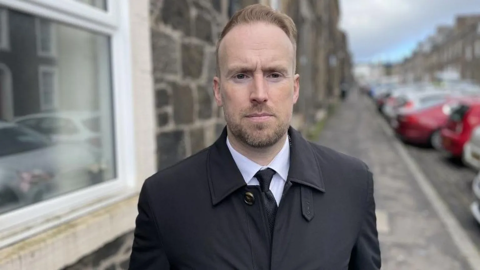 Detective Inspector Forbes Wilson is a young, bearded man with reddish hair. He is wearing a black raincoat, white shirt and black tie and is standing in a street of stone buildings with a line of cars parked facing towards or away from the kerb.