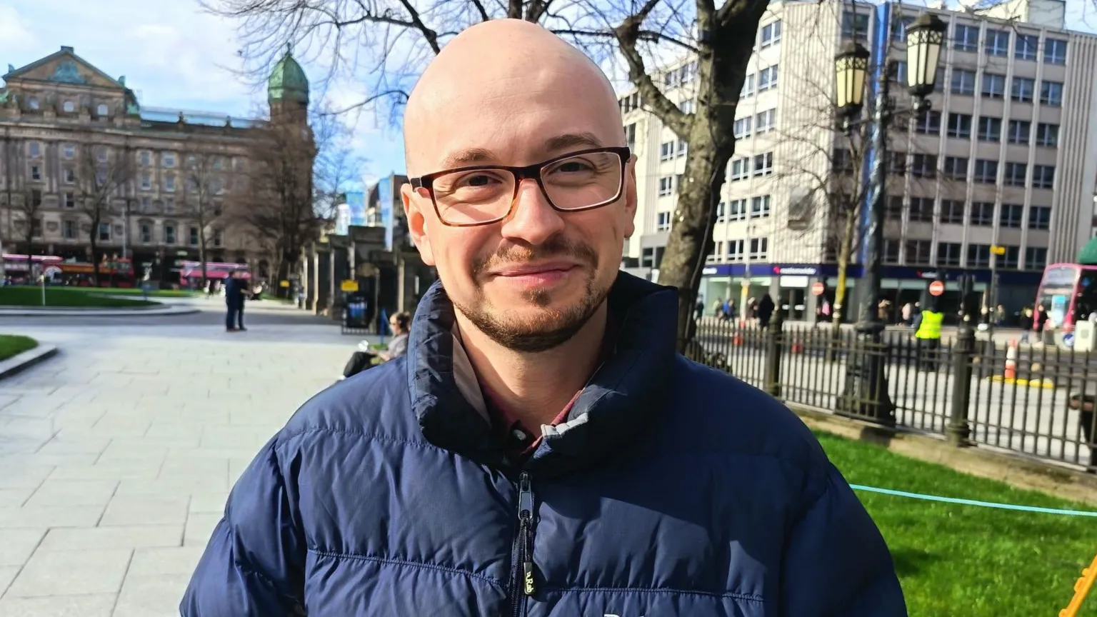 Ben Turner is shaven-headed and is wearing black-framed glasses. He is wearing a dark blue coat and is standing in the grounds of Belfast City Hall