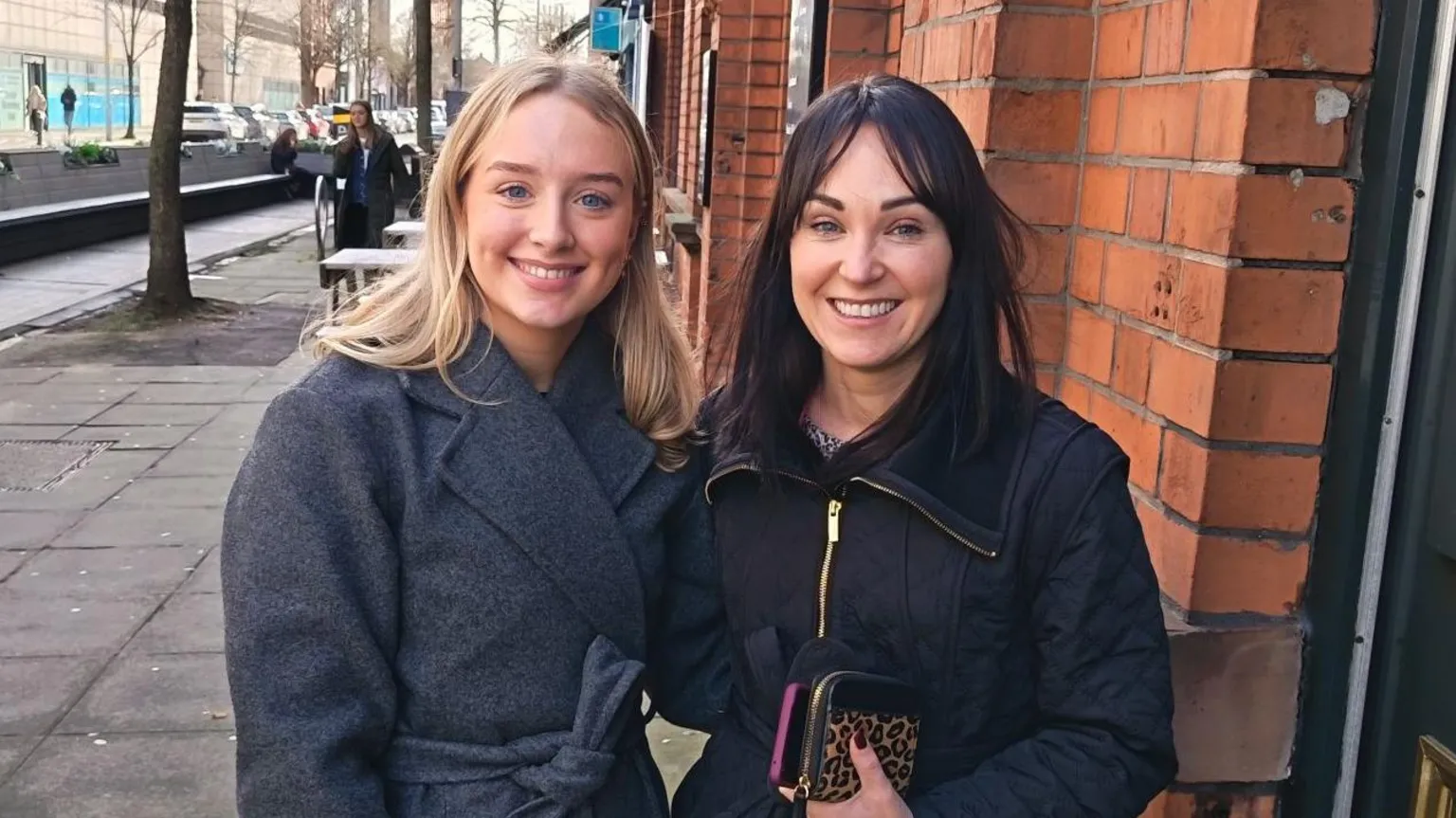 Natalie (on the right) has long dark hair and is smiling. She is wearing a dark coat and holding a purse. Her friend has long blond hair and is wearing a long grey coat