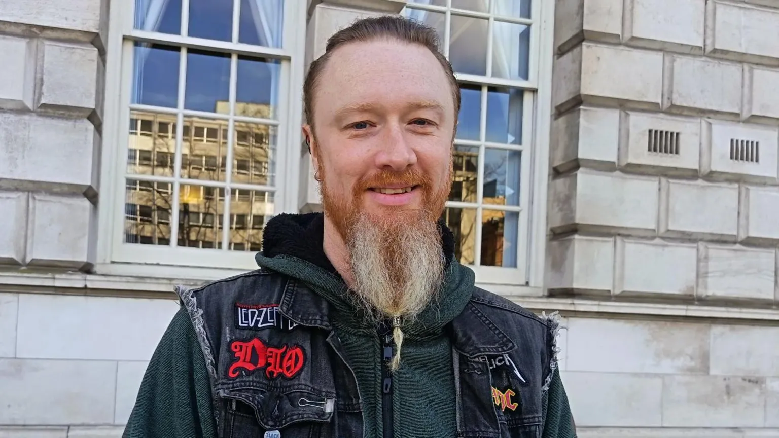 Kenneth McClernon has short brown hair slicked back and a long ginger/grey beard. Her is wearing an armless denim jacket with patches on heavy metal groups on it and a green hoodie