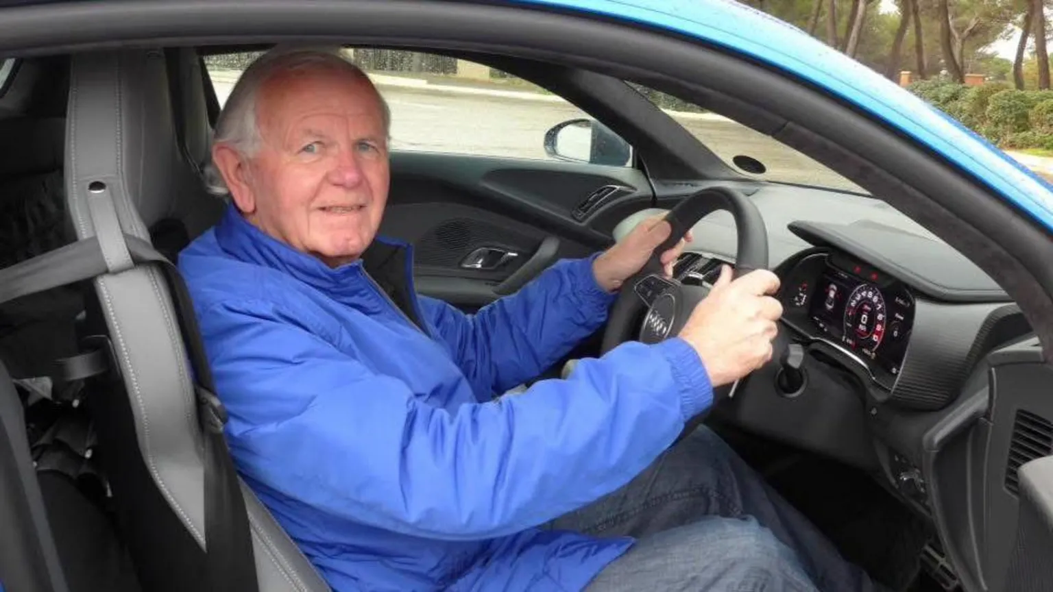 Ian Lynas An older man, with grey hair, sitting in a blue Audi car. The picture is taken from outside the driver side, with the door open. The man is wearing jeans and a blue coat. His hands are on the steering wheel.