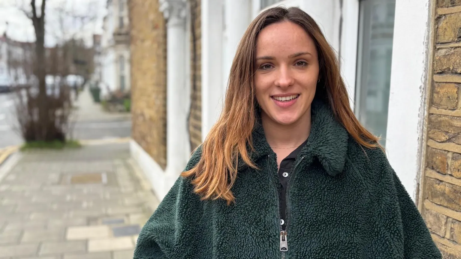 Hazel Shearing / BBC Katie, who has long hair, stands on a residential pavement beside brick and white buildings, wearing a dark green textured zip‑up coat. The street is quiet, with parked cars and bare trees visible in the background.