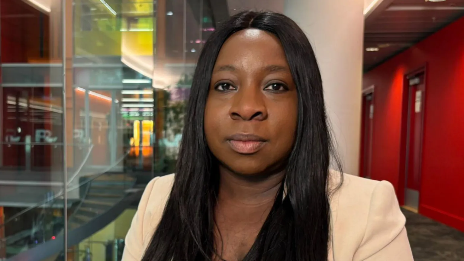 Hayley Clarke / BBC Funmi Olufunwa is seated at a desk in the modern office setting of the BBC building in London. She has long hair, wearing a light-coloured jacket over a dark top. The background shows glass walls, multiple floors and brightly lit interior spaces.