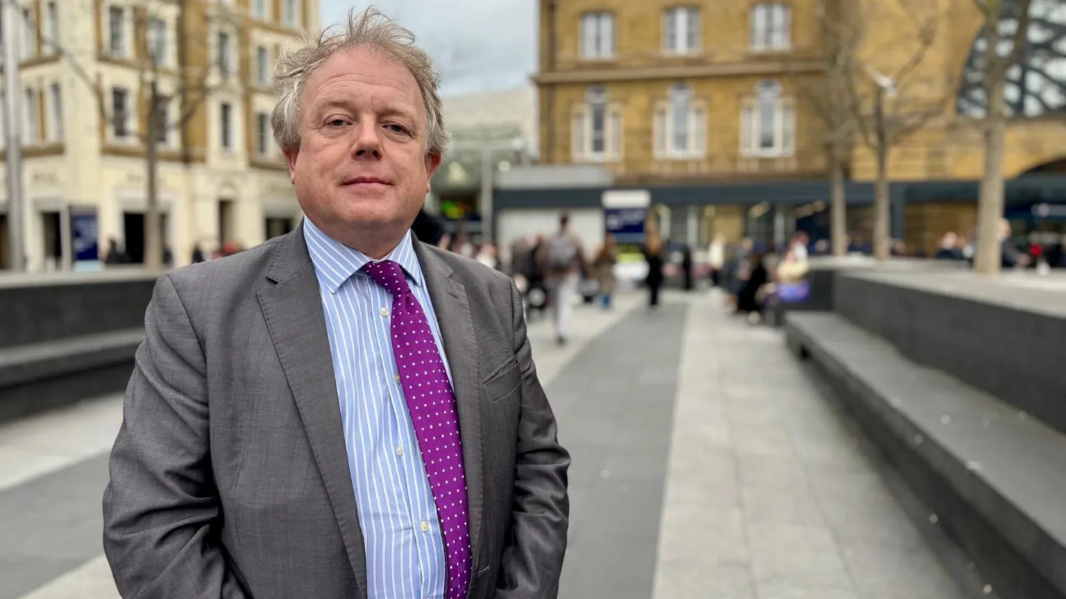 Hazel Shearing / BBC Nick Hillman is wearing in a grey suit and purple spotted tie, standing in an open urban square with stone seating, trees and historic buildings in the background, with people walking and gathering further behind.