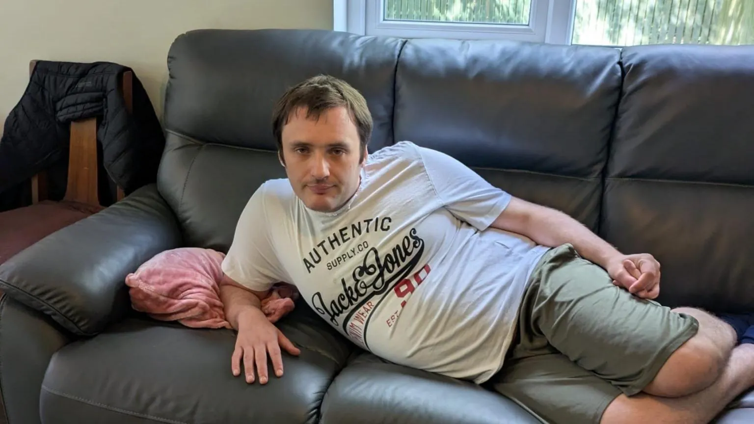 Dawn Jones A man lies on a leather sofa. He is wearing green shorts and a white t-shirt. There is a pink cushion on the sofa. 