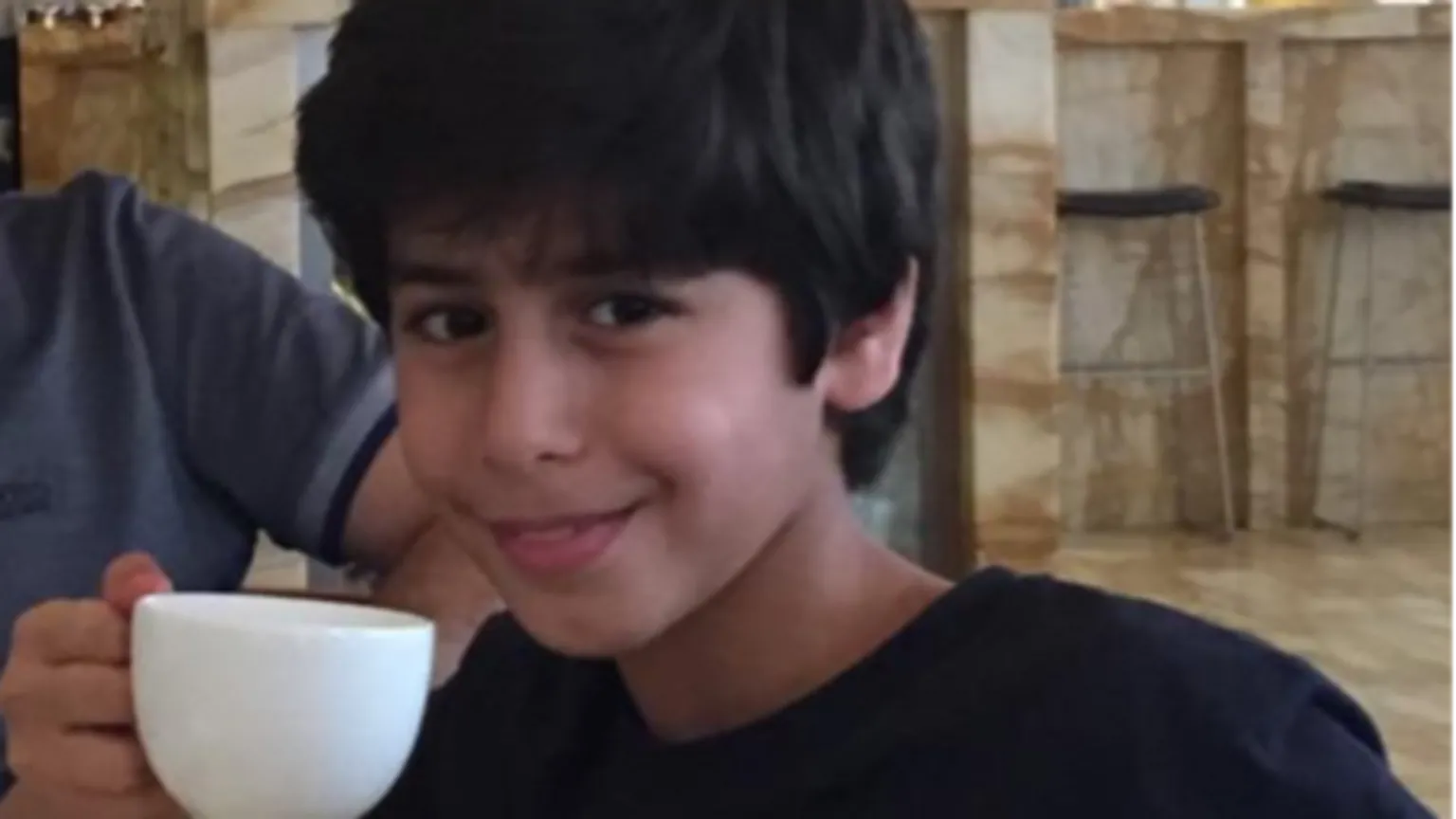 Naif Alqassim Mohammed Algasim pictured as a boy, drinking from a white coffee cup. he is smiling into the camera and wears a black Timberland sweater