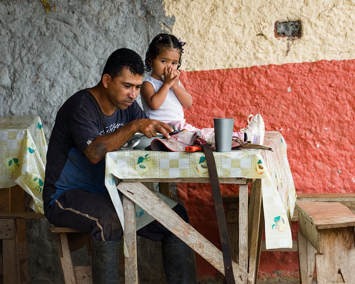 Wilder Gomez, Finca Esperanza’s manager, sits at a simple table eating, with an infant girl sitting on top of the table with him.