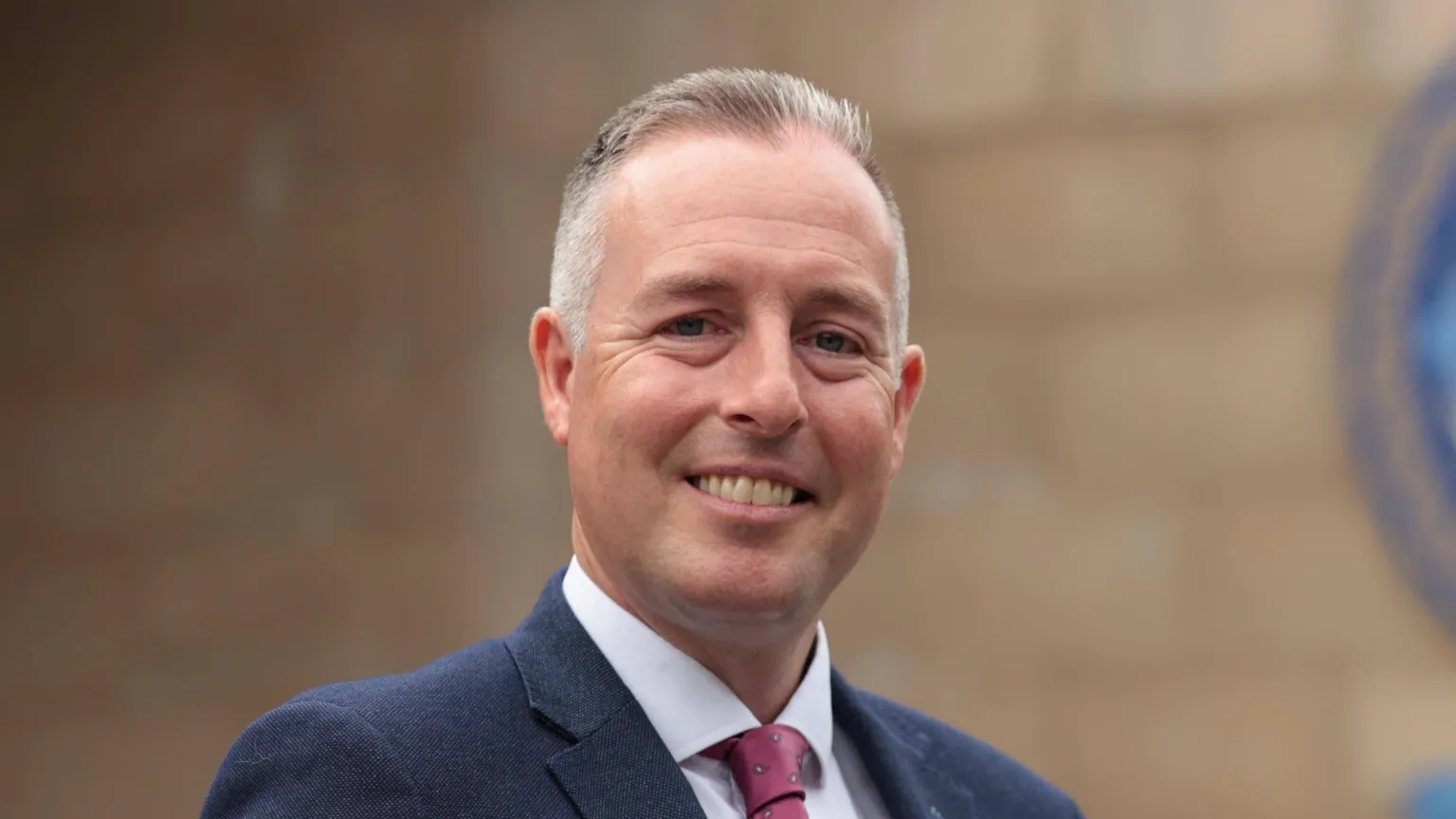  Paul Givan. He has short grey-ish hair, wearing a navy blazer, white shirt and burgundy tie. He is smiling at the camera.
