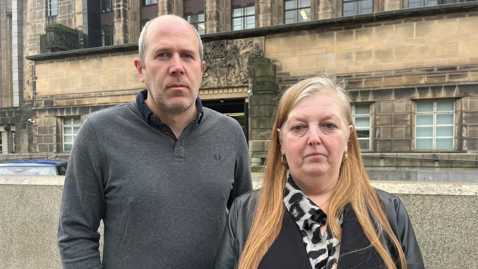 Alistair has short receding hair and stubble. He is wearing a grey jumper. Sharon has long red hair. She is wearing a black jacket.