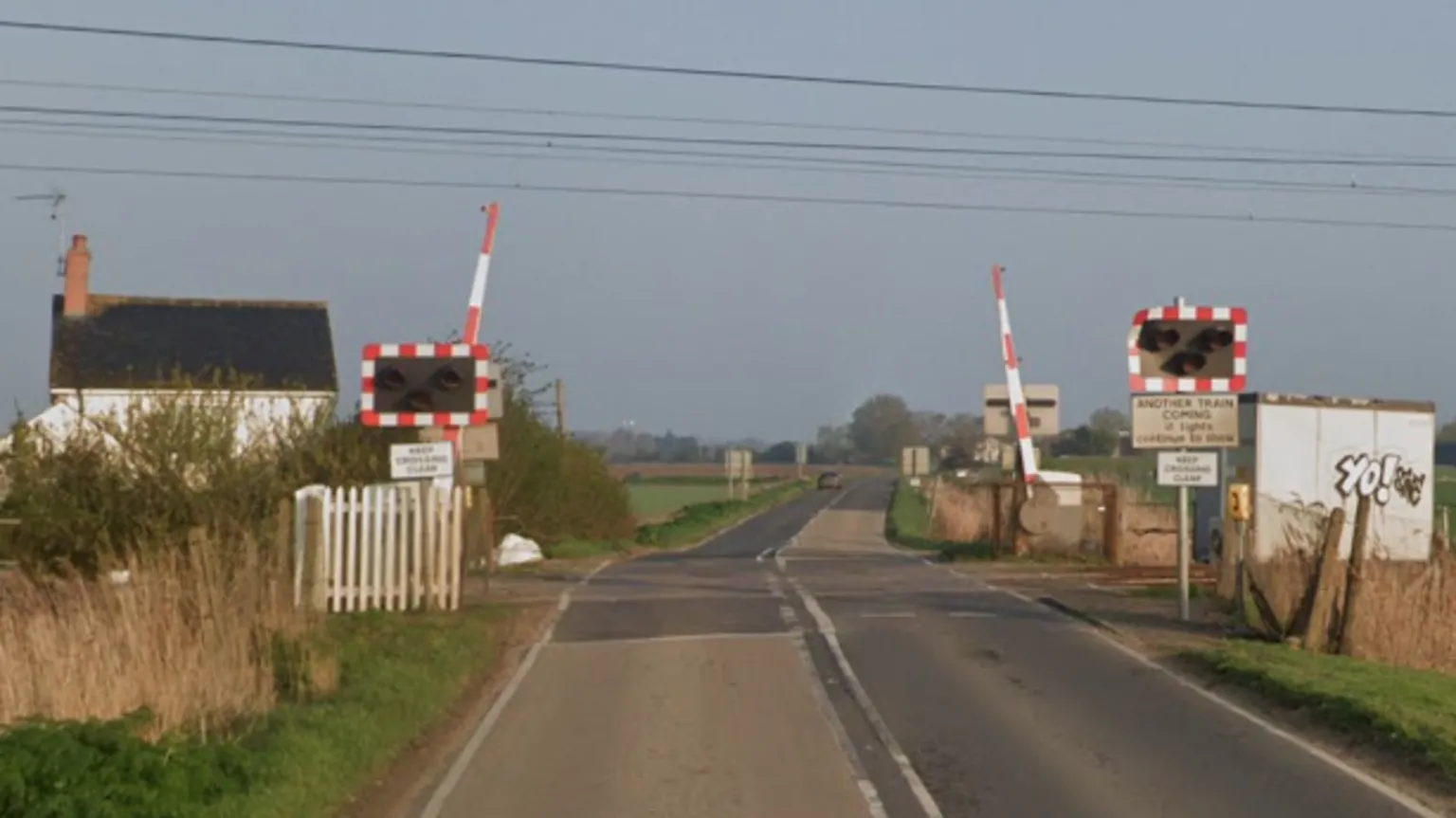 Google A road goes through a level crossing, which is in the middle of fields. There is a small white house on the left and a small hut on the right. There are lights and a barriers, which are open.