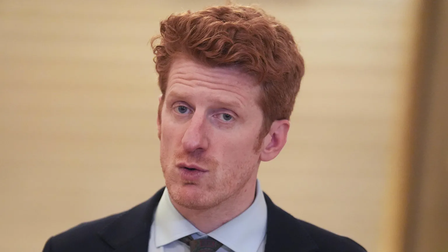  Matthew O'Toole speaking. He has ginger hair and is wearing a navy blazer, a pale blue shirt and a grey tie.