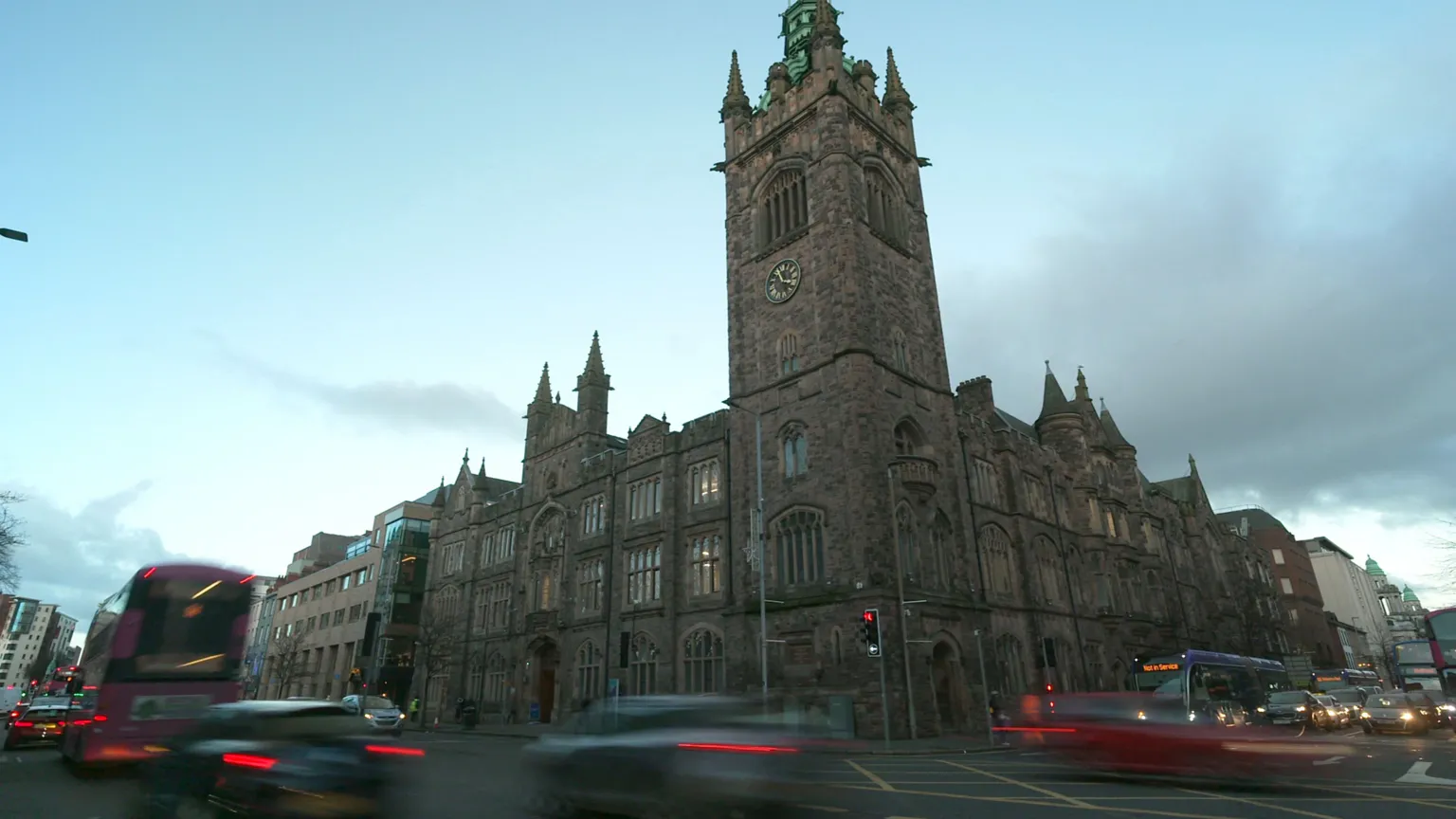 A large church building in Belfast city centre shows, stained glass windows and a bell tower, bearing a gold and black clock. Behind the building is a gloomy, cloudy sky. Traffic is blurred as it passes the camera.