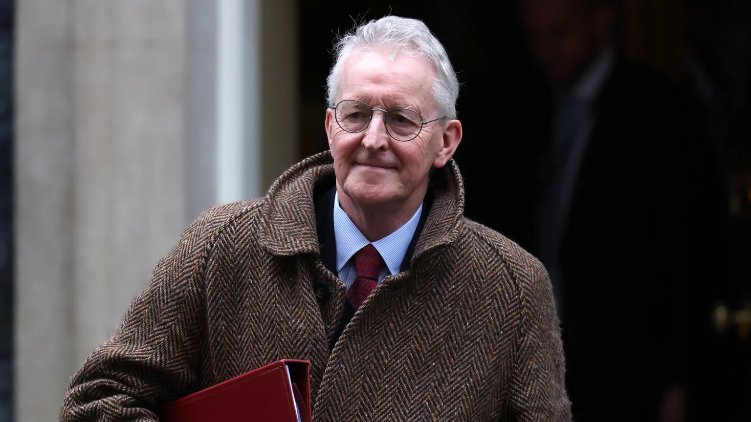 EPA Hilary Benn walking. He is carrying a red file. He is wearing glasses, a brown and cream coat, a blue shirt and a red tie. He has grey hair.