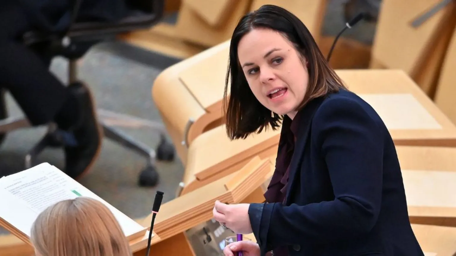 Kate Forbes, who has shoulder length dark hair, speaks in the scottish parliament chamber. She is wearing a dark jacket, and speaking with her head turned back towards her left shoulder