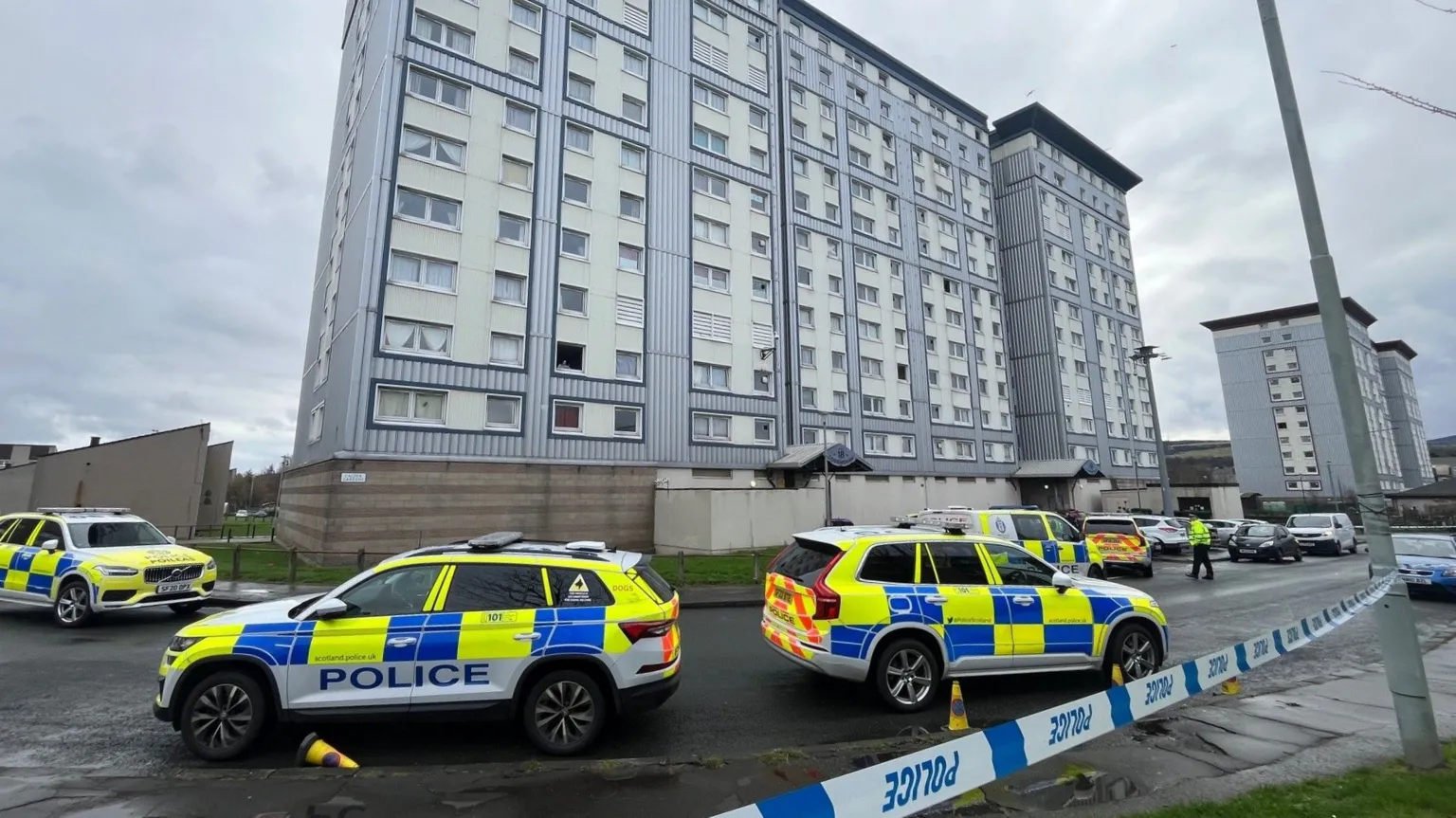 A block of flats clad in blue and white. Police cars line the residential streets and there is police tape blocking off the area, tied to a lamppost.