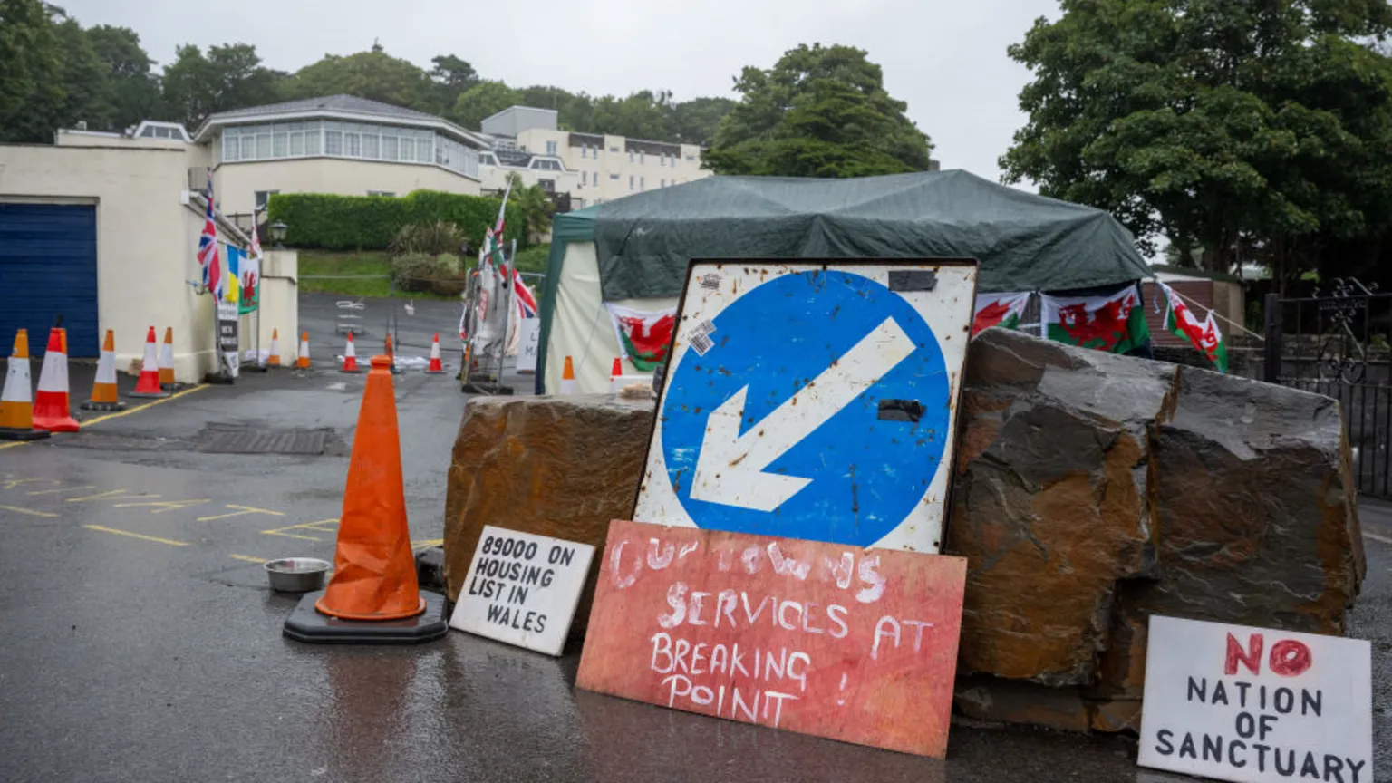  A blockade at the entrance to Stradey Park hotel, with large rocks and orange traffic cones blocking the road. Protest signs lean against the rocks, including messages reading “80000 on housing list in Wales,” “Services at breaking point,” and “No nation of sanctuary.”