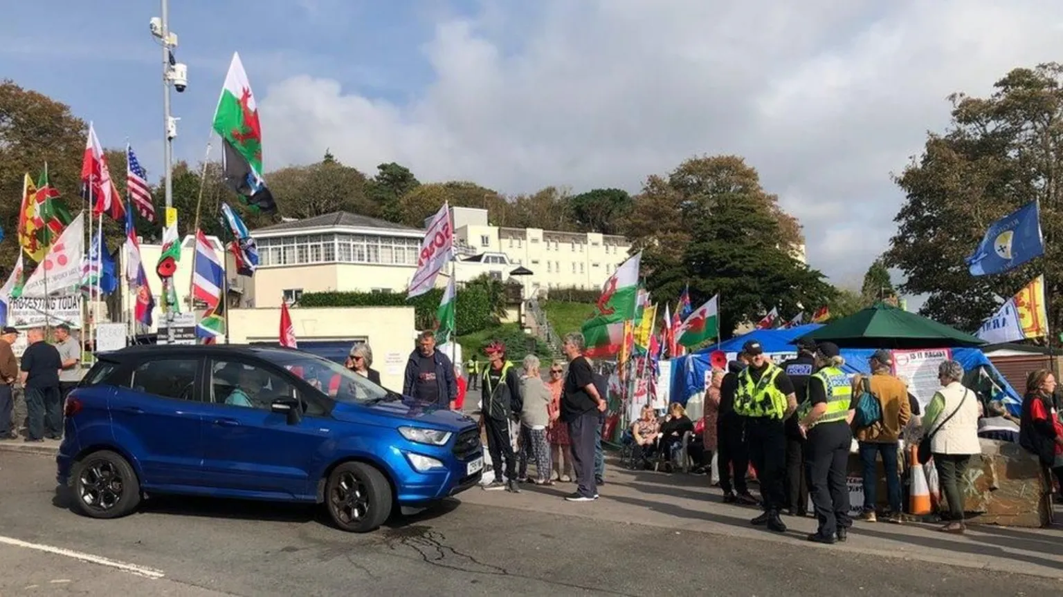  A protest outside the hotel with flags from around the world visible as well as police officers chatting to each other. A dark blue car tries to pass.