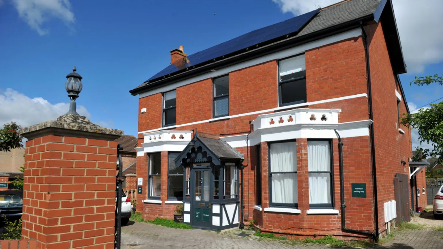 Angie Petkovic A large Victorian or Edwardian style detached suburban house on a sunny day. There are large bay windows on the bottom storey and a Tudor style porch. There are clouds up above. Some cars can be seen parked on the driveway.
