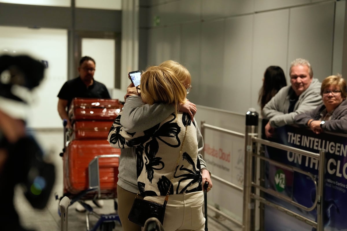 Two women embrace in the middle of an airport terminal in front of a man with a full luggage cart.