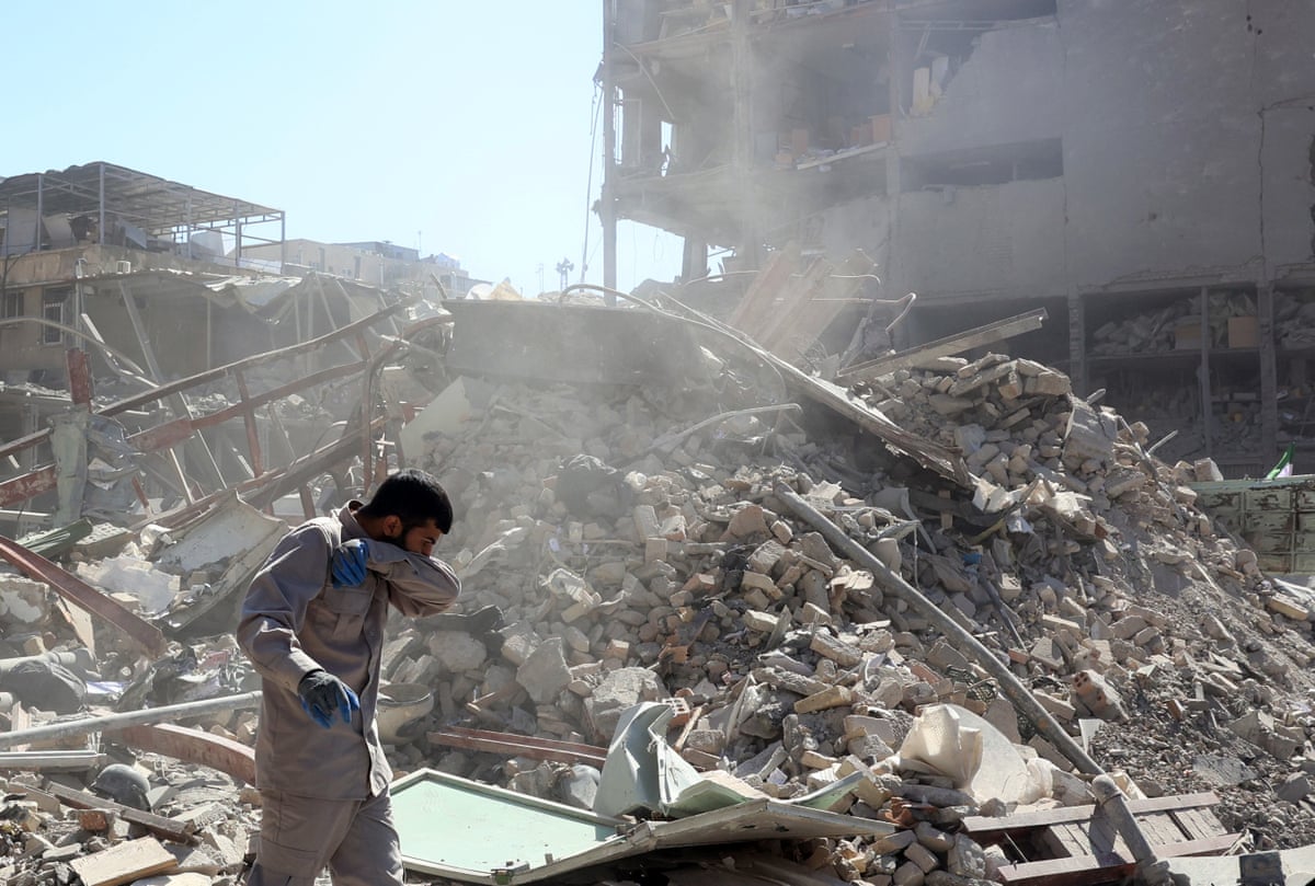 Under a bright sun, a man in a light workers jumpsuit couchs into his arm as dust rises behind him over the debris of a damaged building.