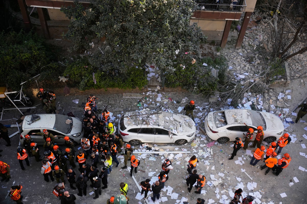 An aerial view into a street shows three parked white sedans covered with dust and debris as officers in bright orange high vis jackets gather in groups to chat. The road is covered in debris and beyond the sedans is a downed tree and some rubble.