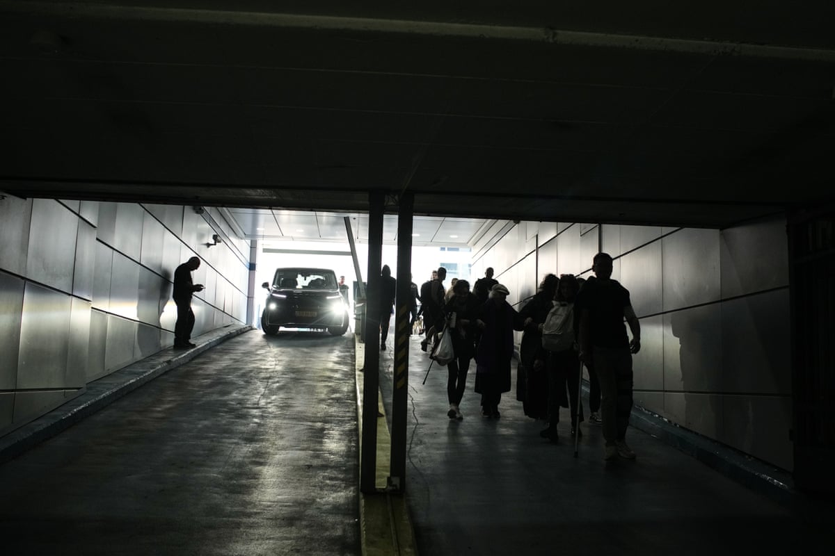 A small crowd of people walk down the ramp of an underground parking lot. Everything is cast in darkness except for the light coming into from the entrance.