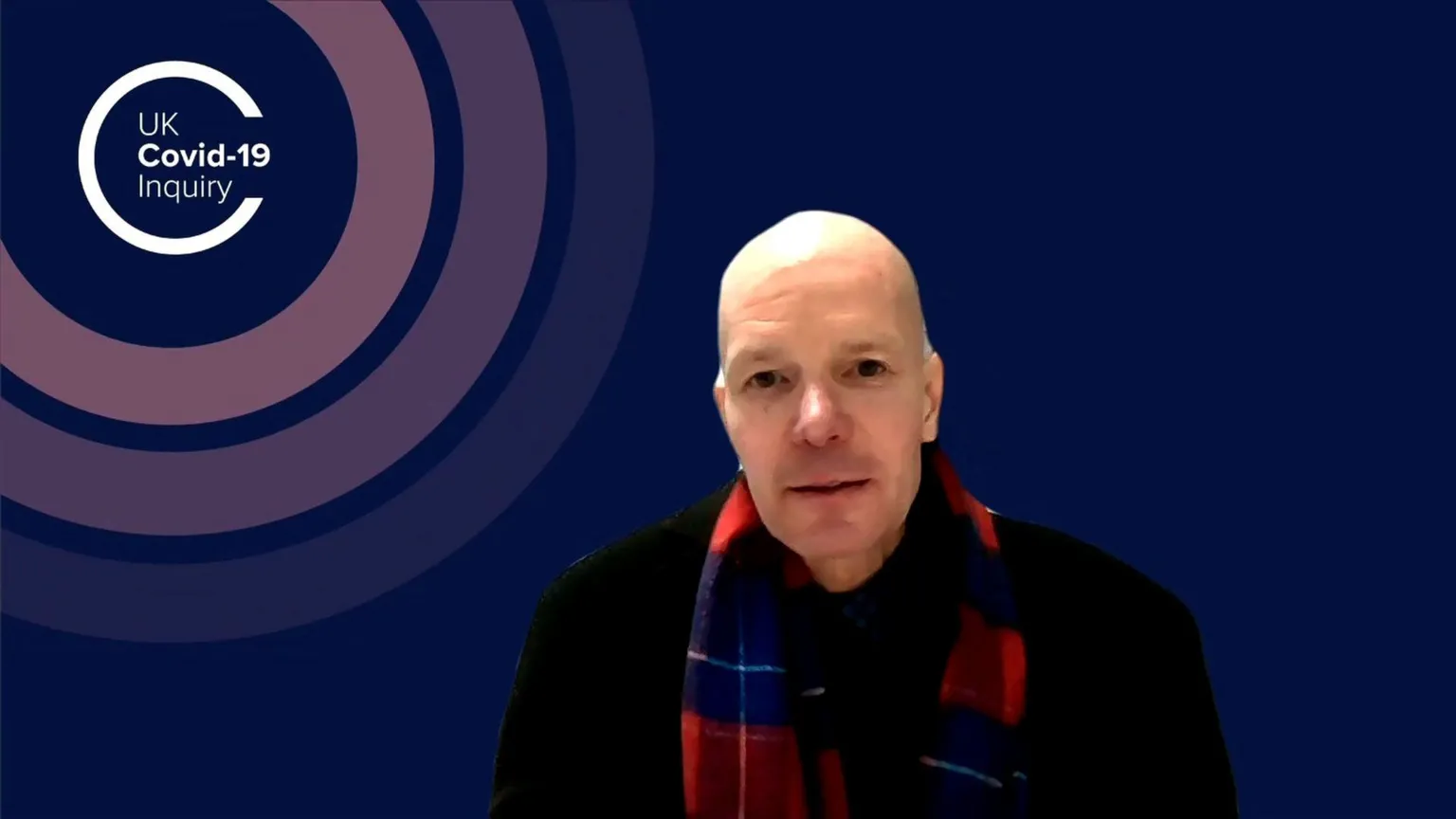 A man is giving in an online meeting. A virtual backdrop states UK Covid-19 Inquiry. He is wearing a red and blue checked scarf and a black jumper.