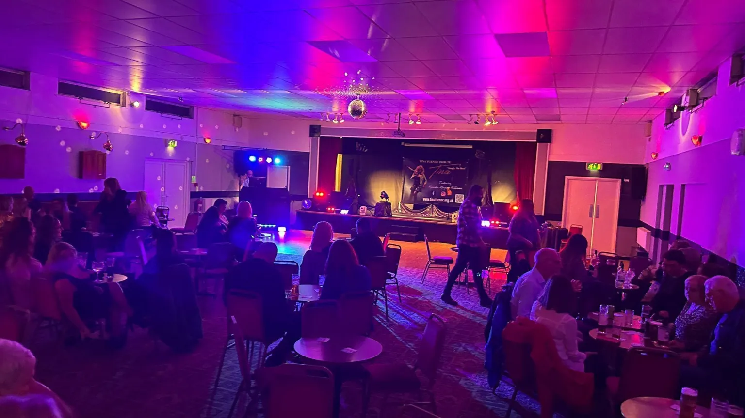 Connah's Quay Labour Club Connah's Quay Labour Club function room, with purple and red hue lights. Round tables with people sat around them face a stage at the back of the hall, with a disco ball attached to the ceiling.