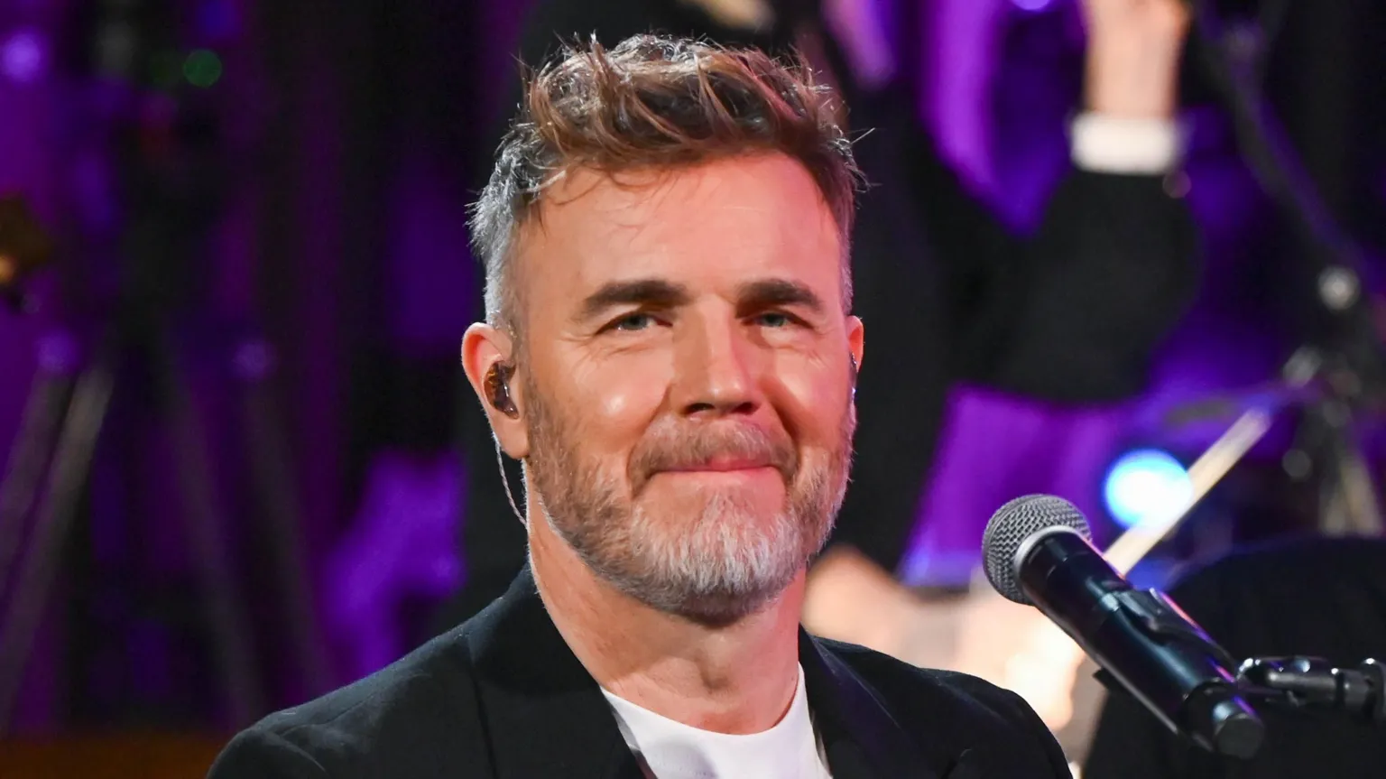 Barlow sat at a piano as an adult. He wears a black jacket and white shirt and has short hair and stubble.