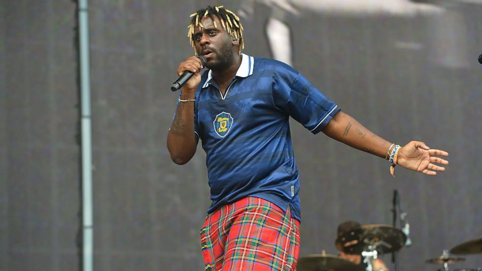 Myles Smith, a singer with blonde dreads, wearing a blue Scotland top and red and green kilt. He is holding a microphone in one hand and has the other outstretched. He is on a stage with a drumkit behind him.