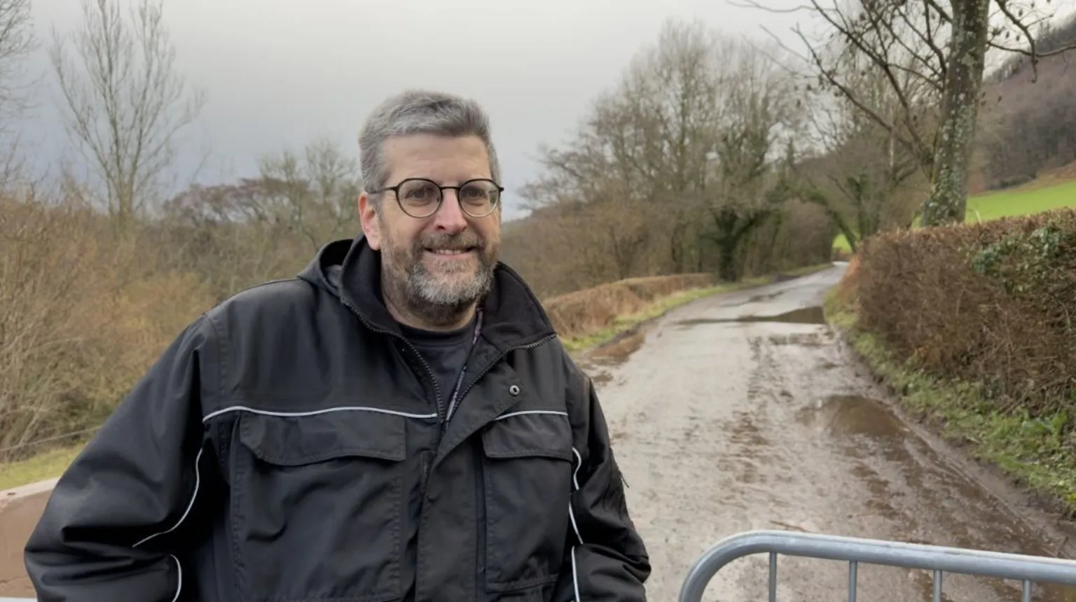 Mark, with short grey hair and a beard, wears a black rain jacket and stands in front of the closed road. He is wearing round glasses and smiles at the camera.