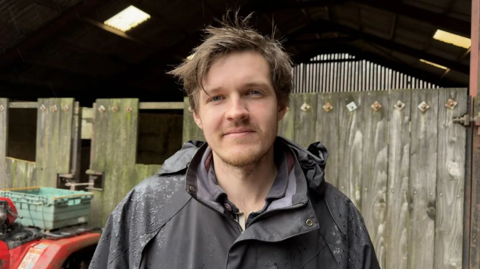 Chris Powell stands in front of a wooden fence. He is wearing a dark jacket specked with rain. He has light brown hair and a short beard.