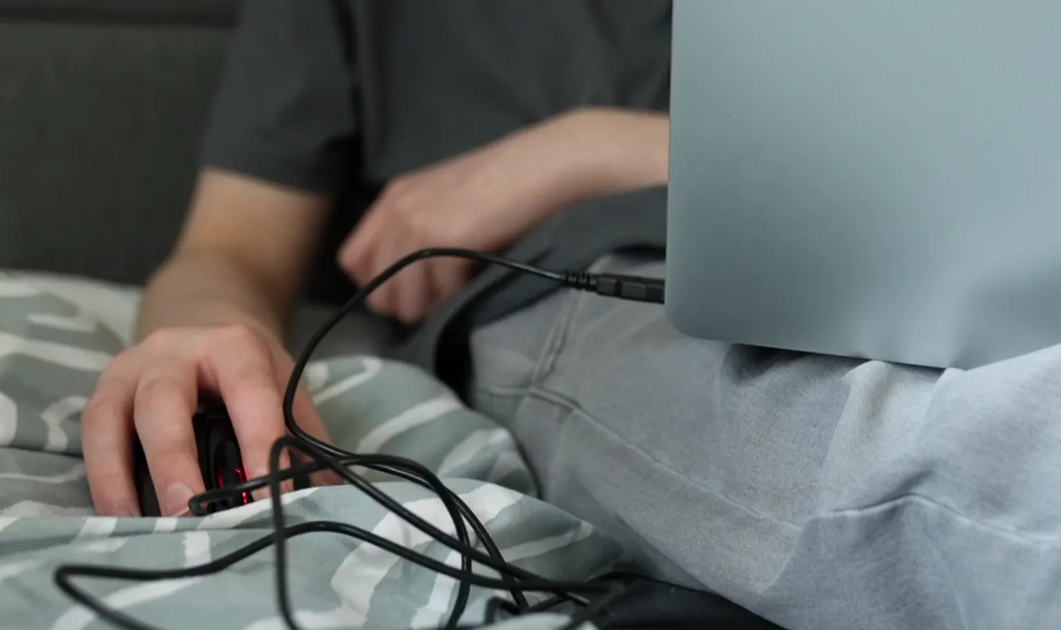 A persons hand on a black mouse as they sit on a turquoise duvet with a laptop on their knee. 
