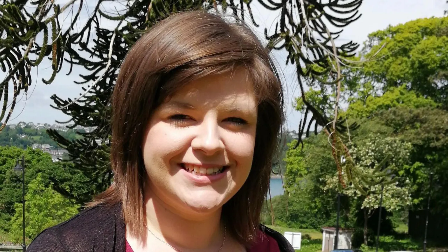 Bangor University A brunette woman wearing a red top and black cardigan, stood in front of a patch of greenery.