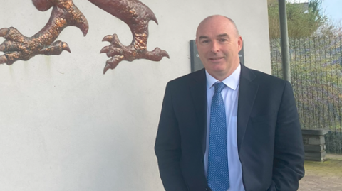 Dyn mewn siwt las a thei glas yn sefyll ar bwys wal wen gyda gwaelod arwydd yr ysgol mewn golwg