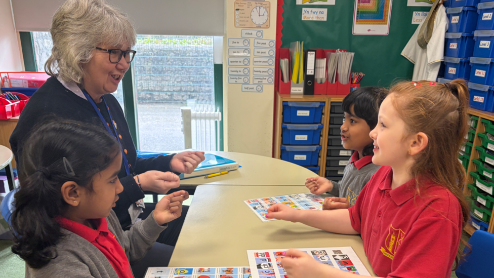 Athrawes a thair merch mewn gwisg ysgol yn eistedd o gwmpas bwrdd gyda cherdiau lliwgar o'u blaenau