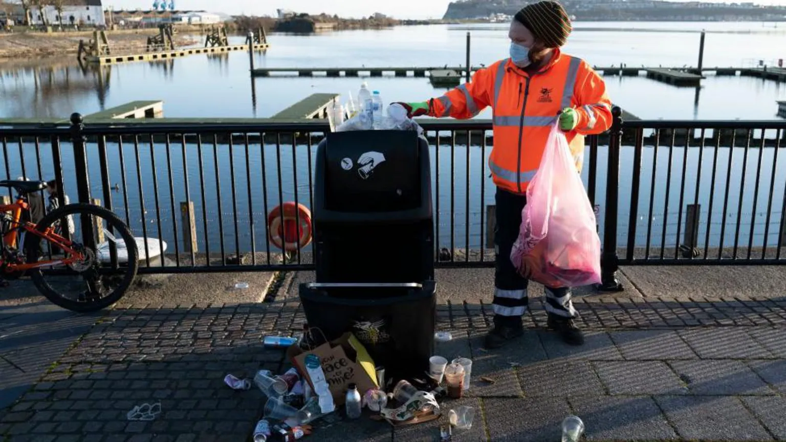 A refuse worker in orange hi-vis holds a clear plastic binbag and empties a bin at Cardiff Bay. The water is behind him.