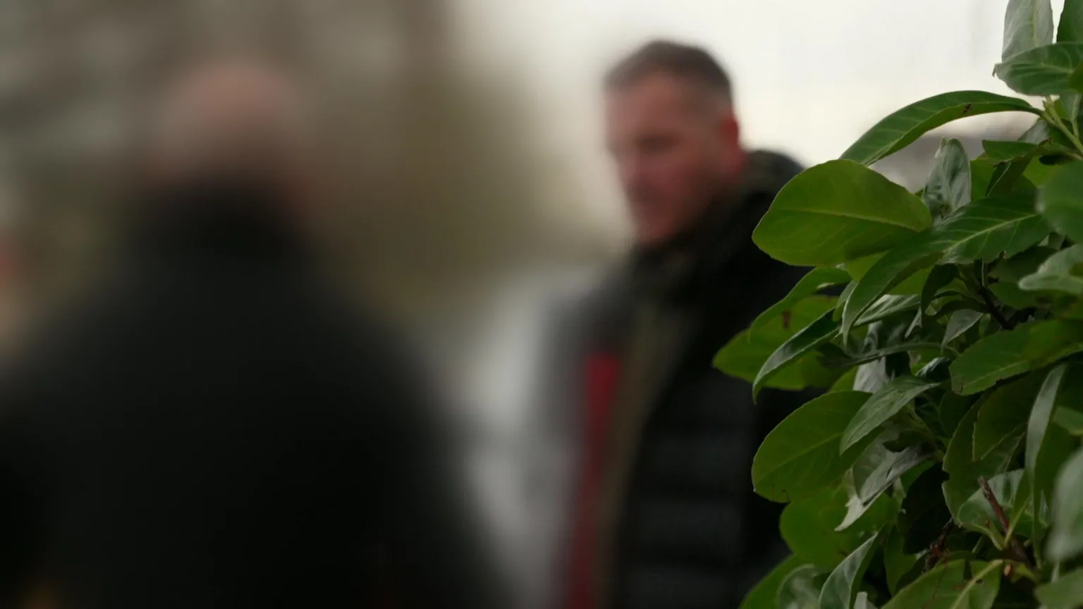 A blurred image of a man talking to another man outside with the leaves of a bush seen in the foreground.