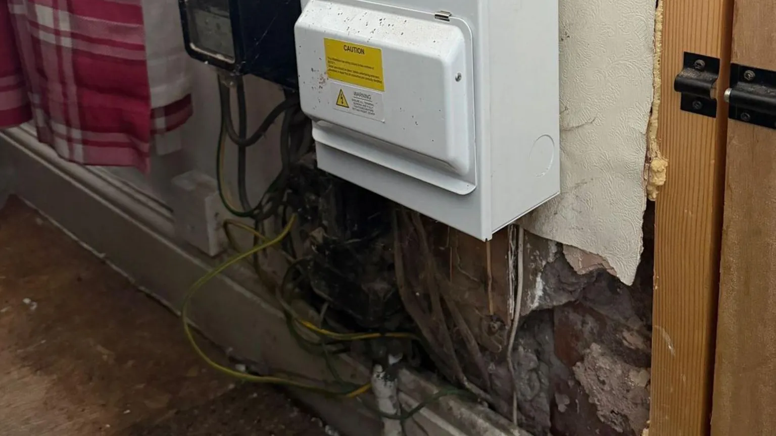 Electric meter fixed to wall with exposed live wiring hanging down underneath and shoddy plasterwork.