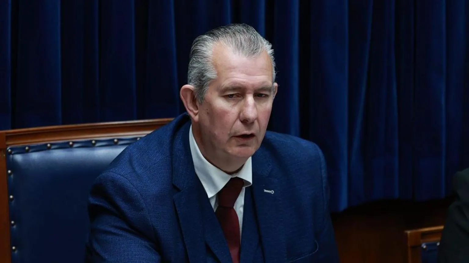 Edwin Poots is wearing a blue suit jacket, red tie and white shirt. He is sitting on the speaker's seat. He has grey, receding hair.
