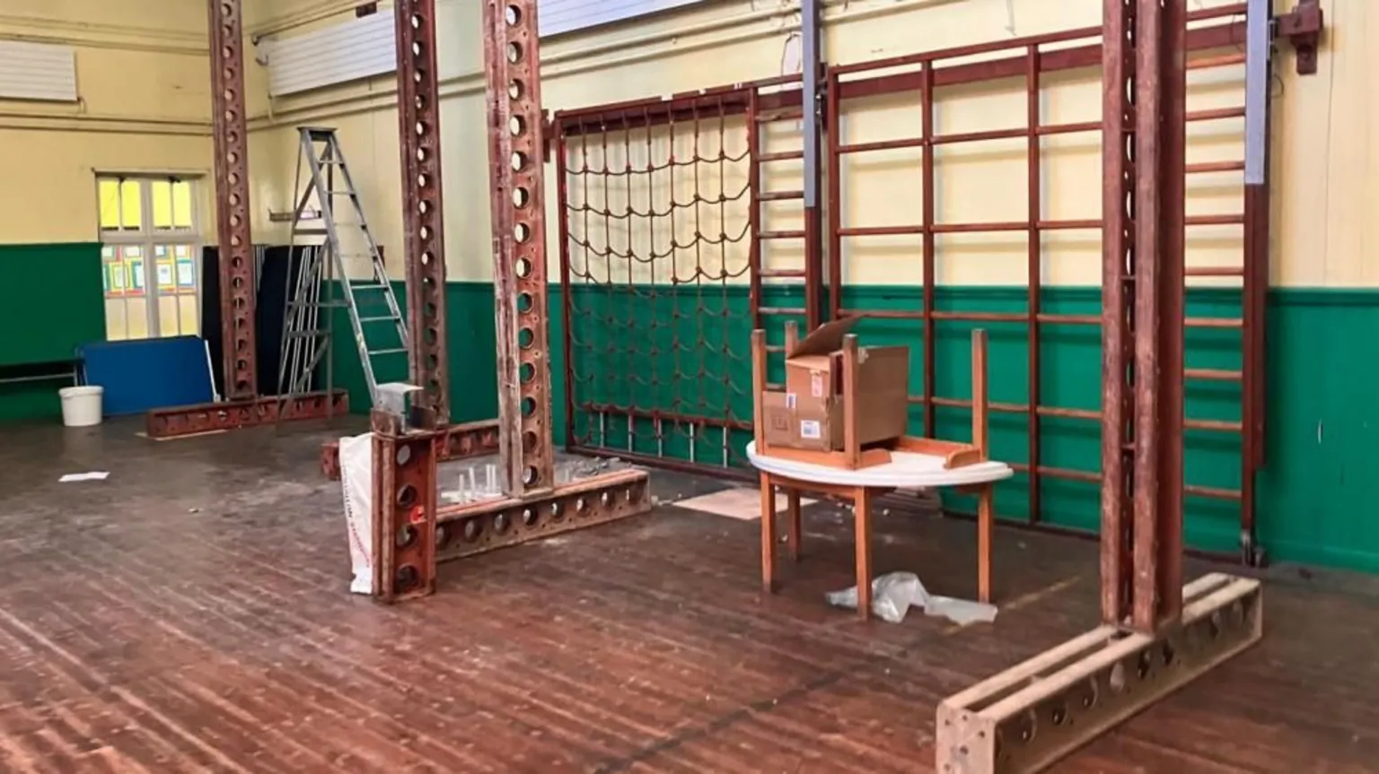 Rosetta Primary School Four tall rusted metal beams are stacked from the floor to help support the roof of a green and white school assembly hall. The dark wooden floor is dusty and there are two tables stacked on top of one another.