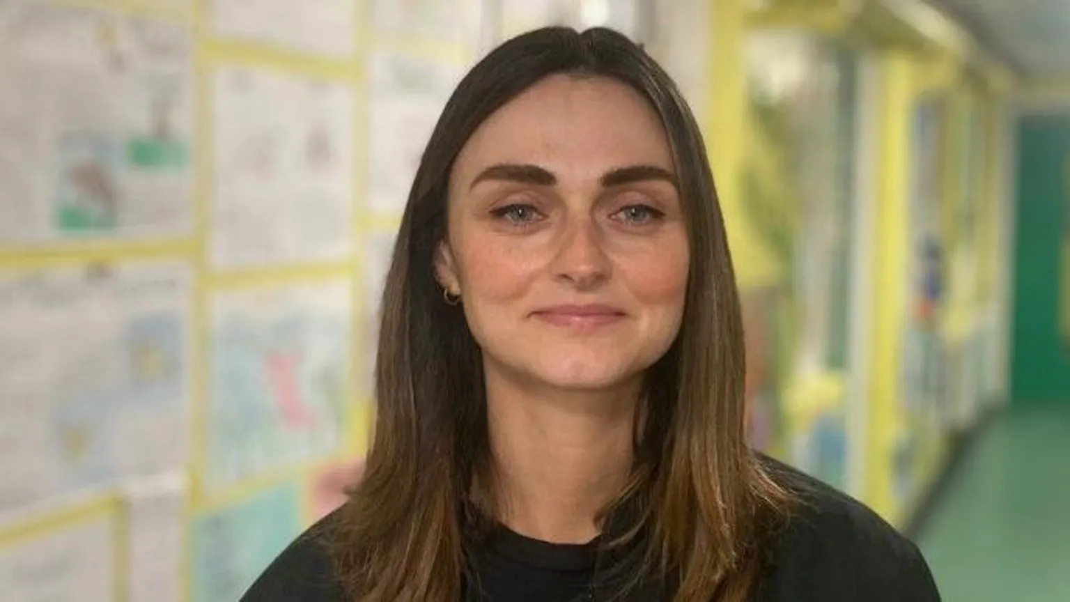 A woman with long dark hair stares into the camera. She is wearing a dark top and is standing in a school corridor.