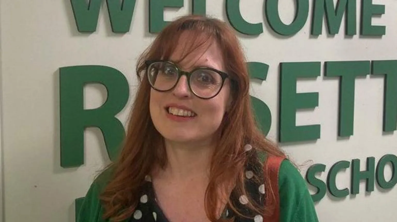 A woman with red hair and big, black glasses stands in front of a sign saying 'Welcome to Rosetta Primary School' in green letters. She is wearing a polka dot blouse and a green cardigan. The brown strap of a handbag is visible over her left shoulder.