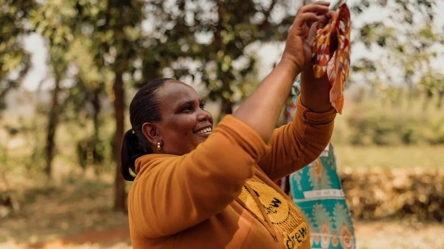 World Vision UK A woman in Tanzania stands outdoors beneath the shade of tall trees, holding her arms up as she clips a freshly washed reusable period pad on to a clothes line. She wears a mustard‑coloured sweater, and the sunlight filters softly through the leaves around her. Behind her, the ground is covered in dry grass and scattered foliage, with a backdrop of green vegetation stretching into the distance.